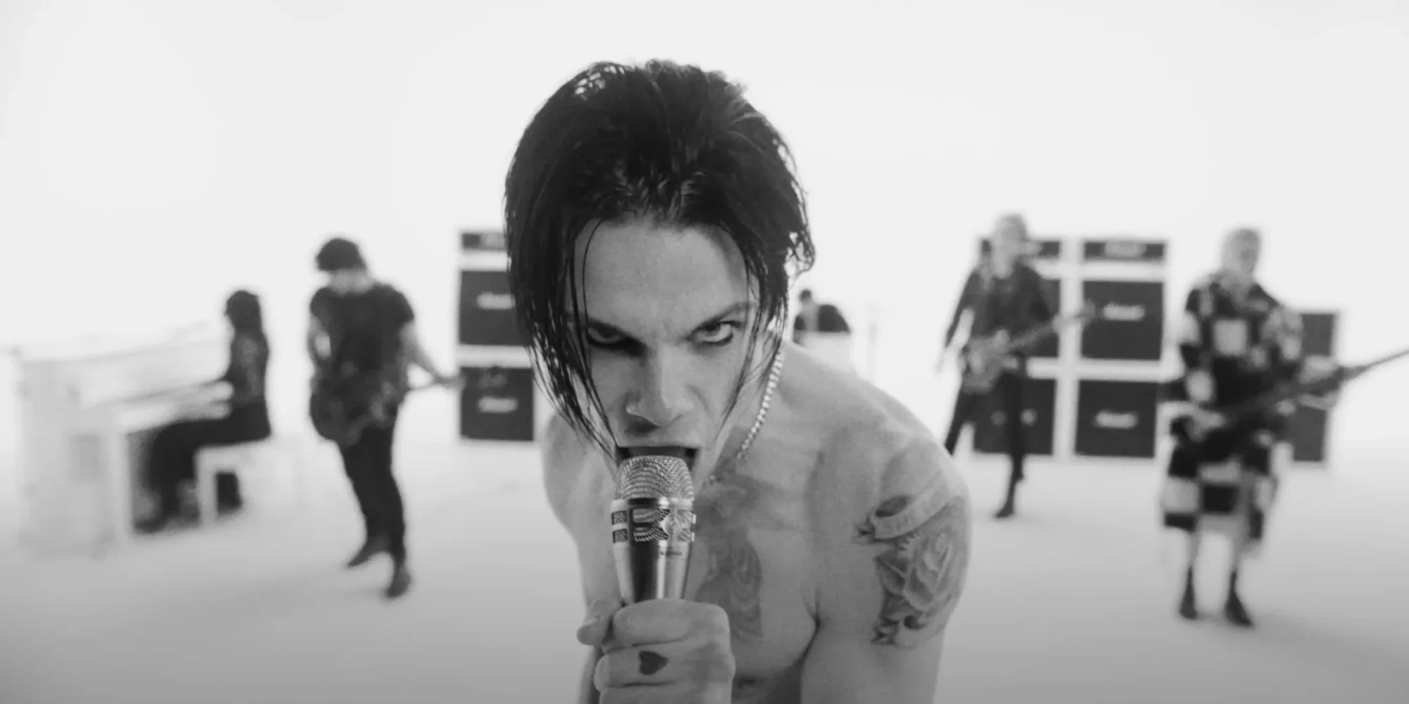 Singer with tattoos gripping mic intensely, band playing in a white studio. Amplifiers stacked in the background. Monochrome image.