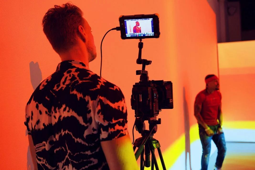 Photographer in a patterned shirt films a person in red against an orange-lit backdrop. Camera screen shows a preview. Vibrant mood.