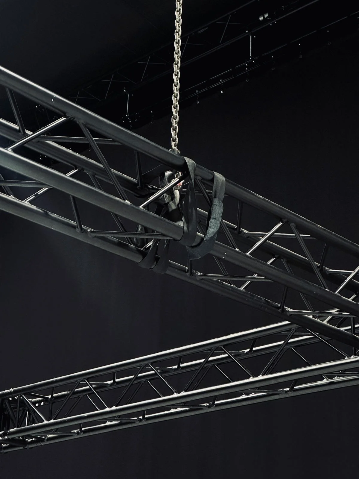 Metal trusses suspended by chains in a dark, industrial setting. The mood is utilitarian, with a focus on structural elements.