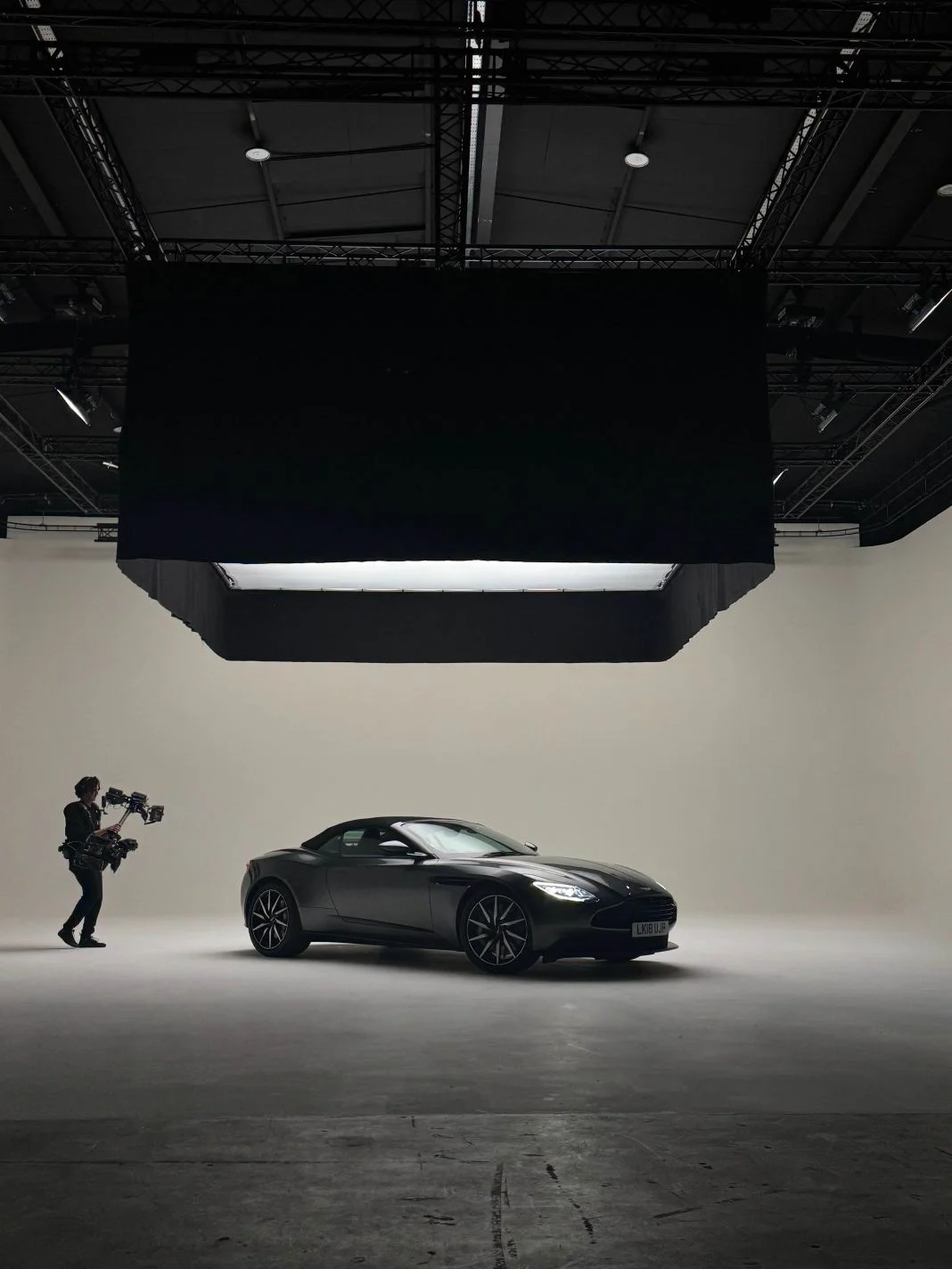 Camera operator films a sleek black sports car in a spacious, well-lit studio. Multi-level ceiling and a large light fixture above.