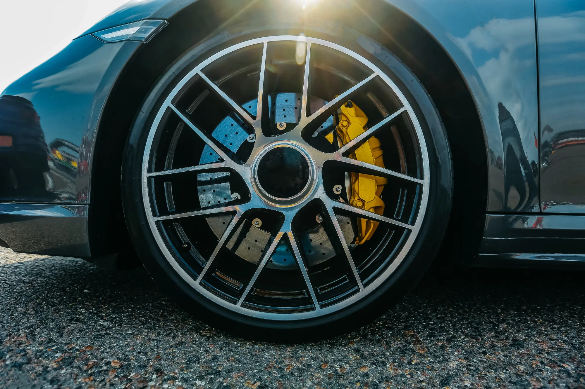 Close-up of a car wheel with a yellow brake caliper, shiny black rim, sunlight glare, on a textured asphalt surface.