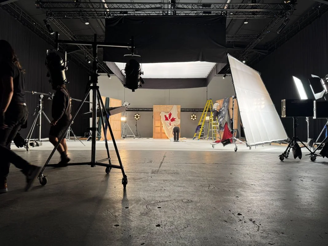 Film studio set with crew members adjusting equipment. A draped Canadian flag is visible. Bright lights and ladders are in the background.