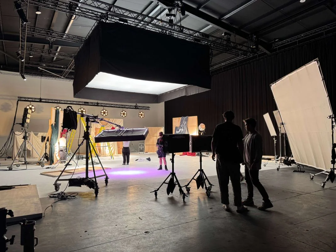 Film studio with lighting equipment and crew setting up a scene. Two people stand in silhouette. Bright lights illuminate backdrops.