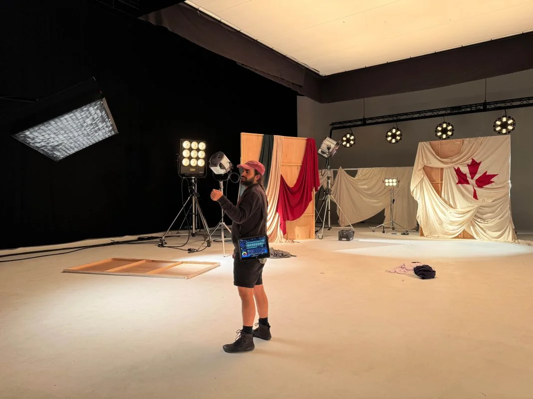 Man in a studio with lighting equipment, wearing a red cap. Backdrop features a maple leaf and draped fabric, creating a creative setting.