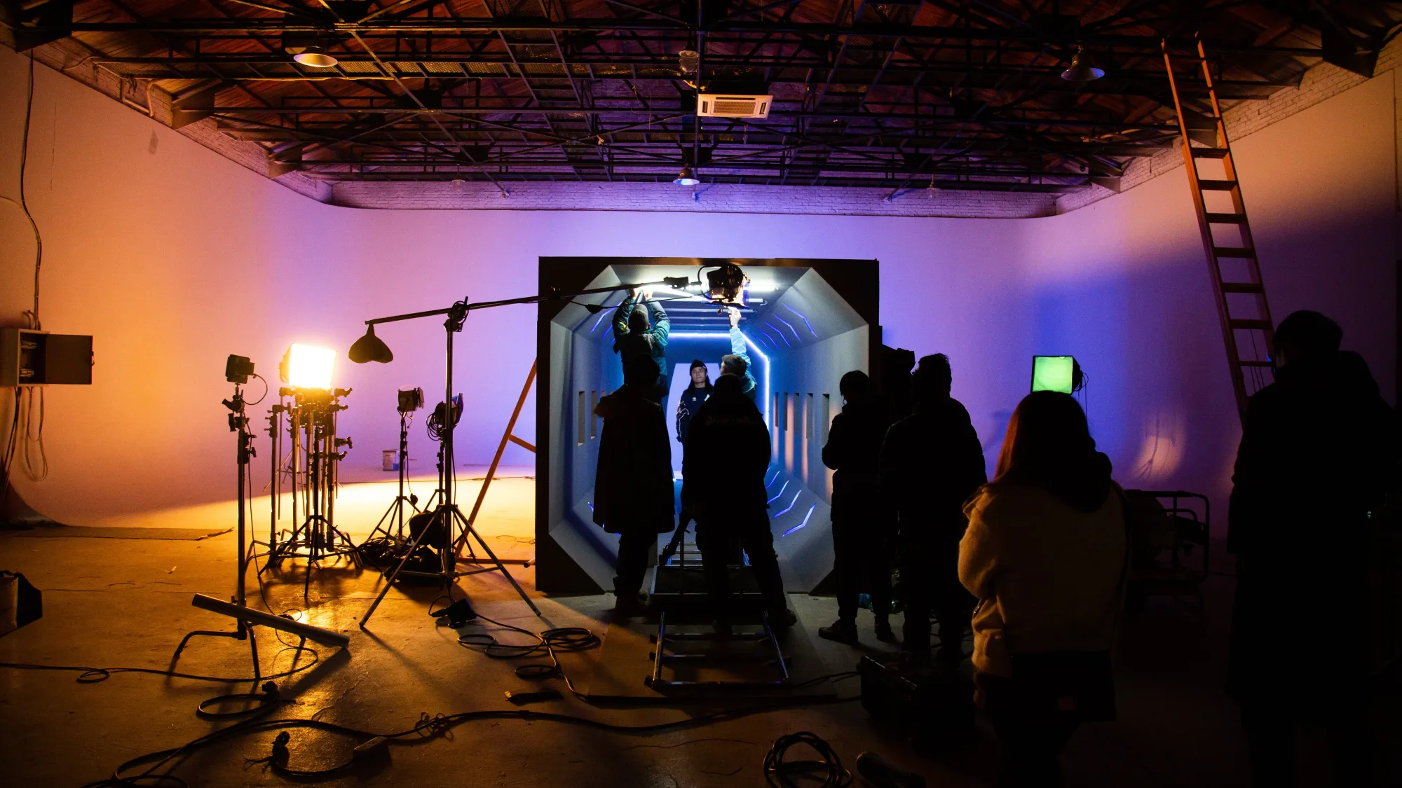 Film set with crew adjusting lights in a dimly lit studio. A tunnel set glows blue, surrounded by equipment. Orange and purple hues dominate.
