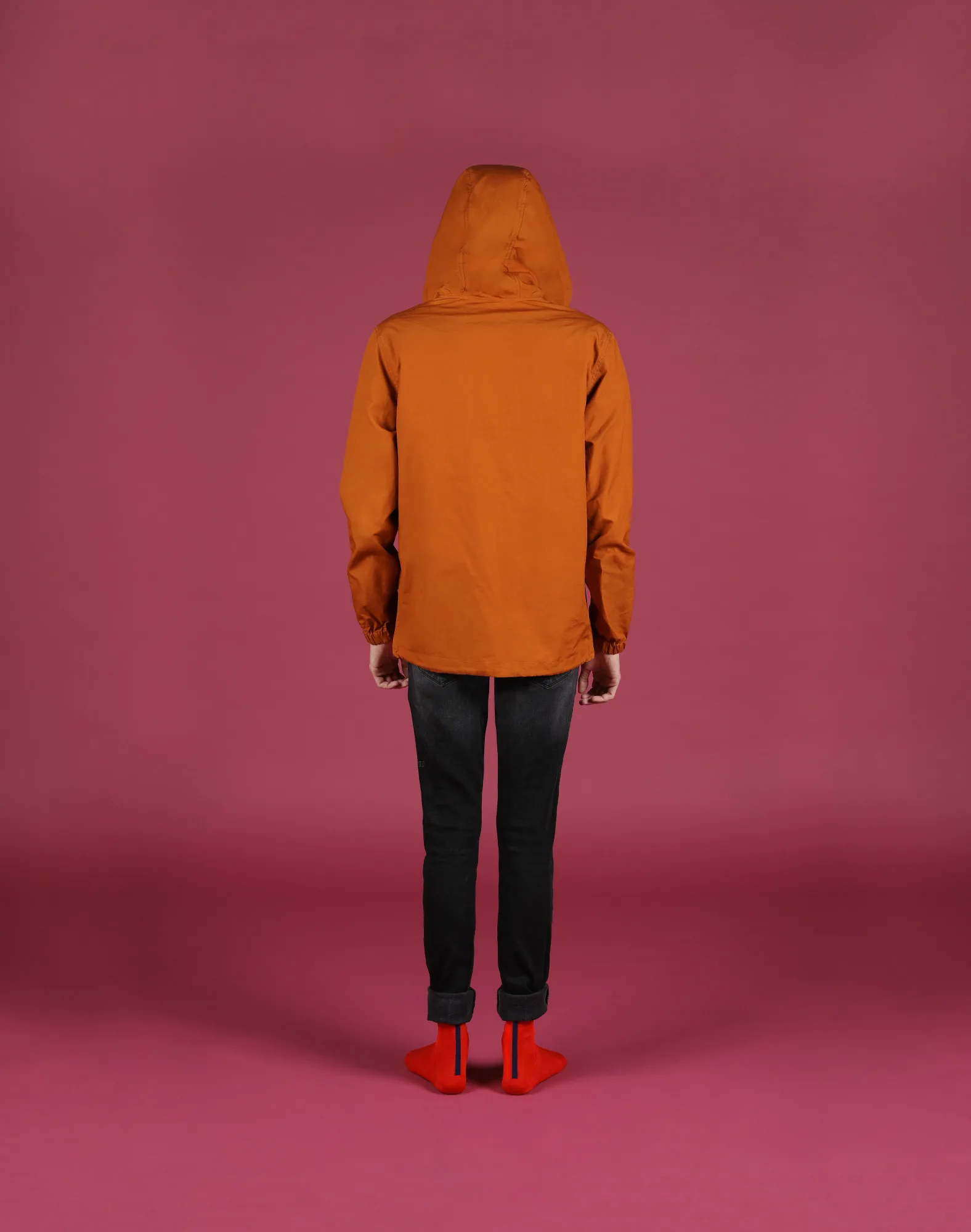 Person in orange jacket and red shoes faces a pink wall, standing still. Background and person have vibrant, contrasting colors.