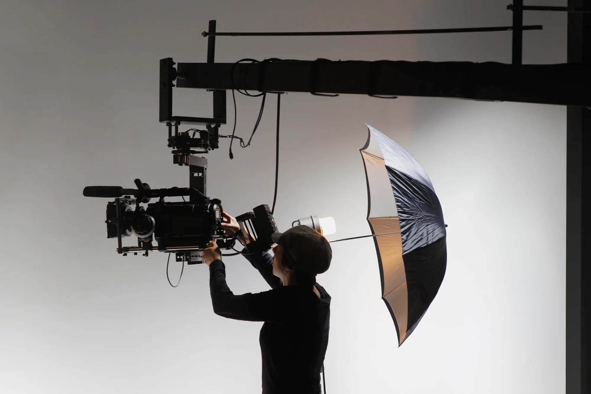 Person adjusts a large camera on a crane, under studio lighting. An umbrella softens the light. Background is plain white.
