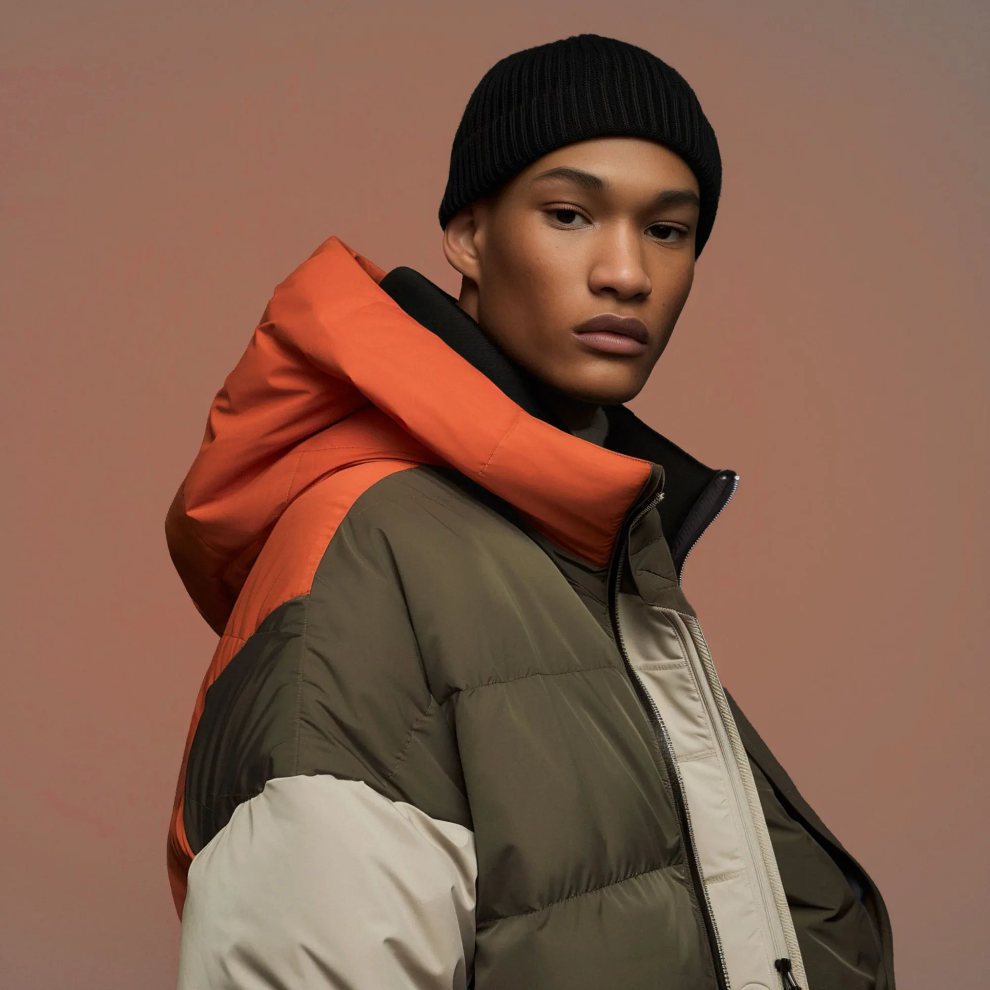 Person in a color-blocked puffer jacket and black knit beanie against a soft brown background, looking serious and composed.