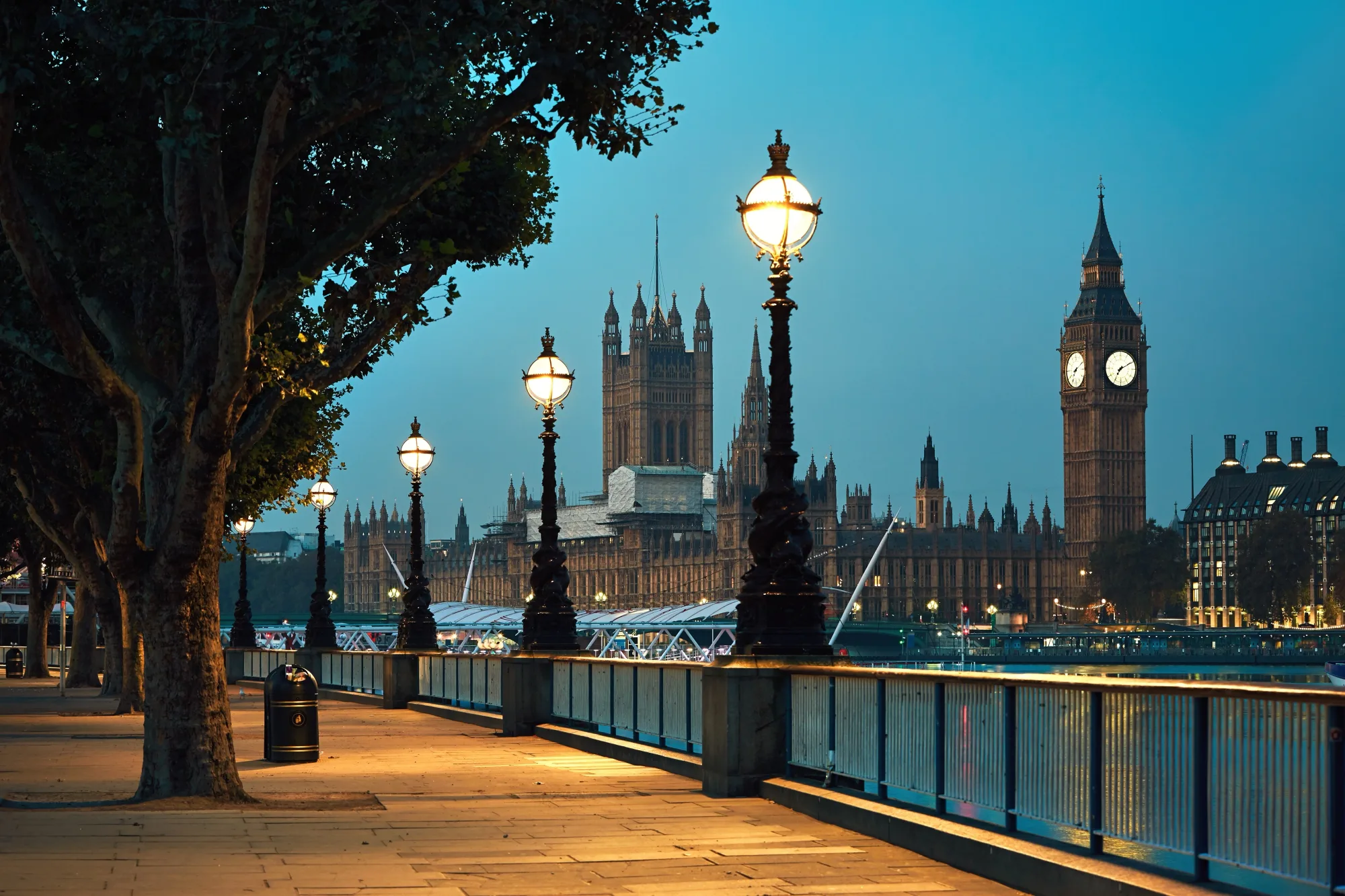 Pathway along the Thames with lit street lamps at dusk. Big Ben and Parliament in view, creating a serene and historic atmosphere.