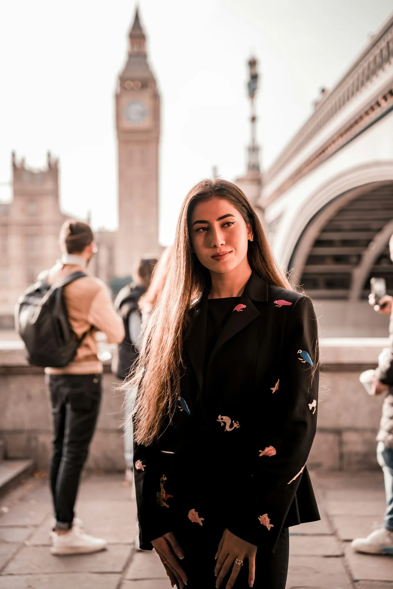 Woman in a black jacket with colorful patterns stands in front of Big Ben, with tourists and a bridge in the background. Mellow lighting.