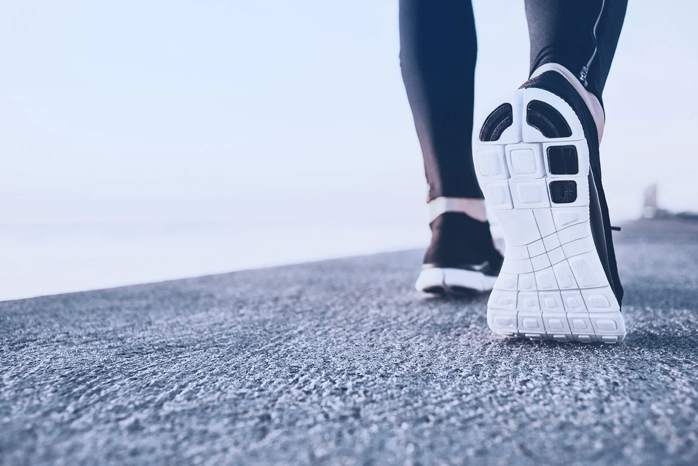 Close-up of a person in black leggings walking on a sunny, textured path by the sea. Focus on white sneaker soles against a clear sky.