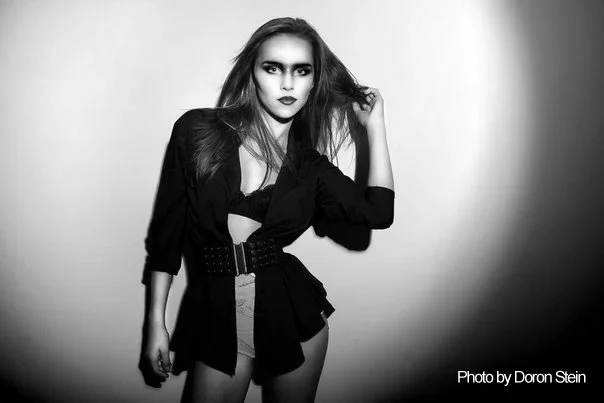Woman in dramatic black outfit poses against a light background. Bold makeup, confident expression. Text: "Photo by Doron Stein."