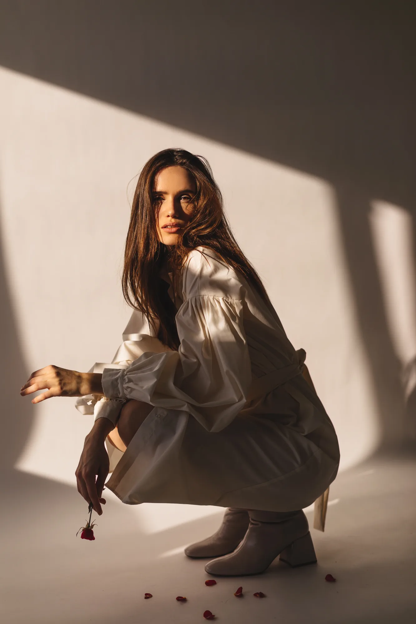 Woman in a white dress crouches in sunlight holding a red rose. Petals scattered on the floor, creating a serene, reflective mood.