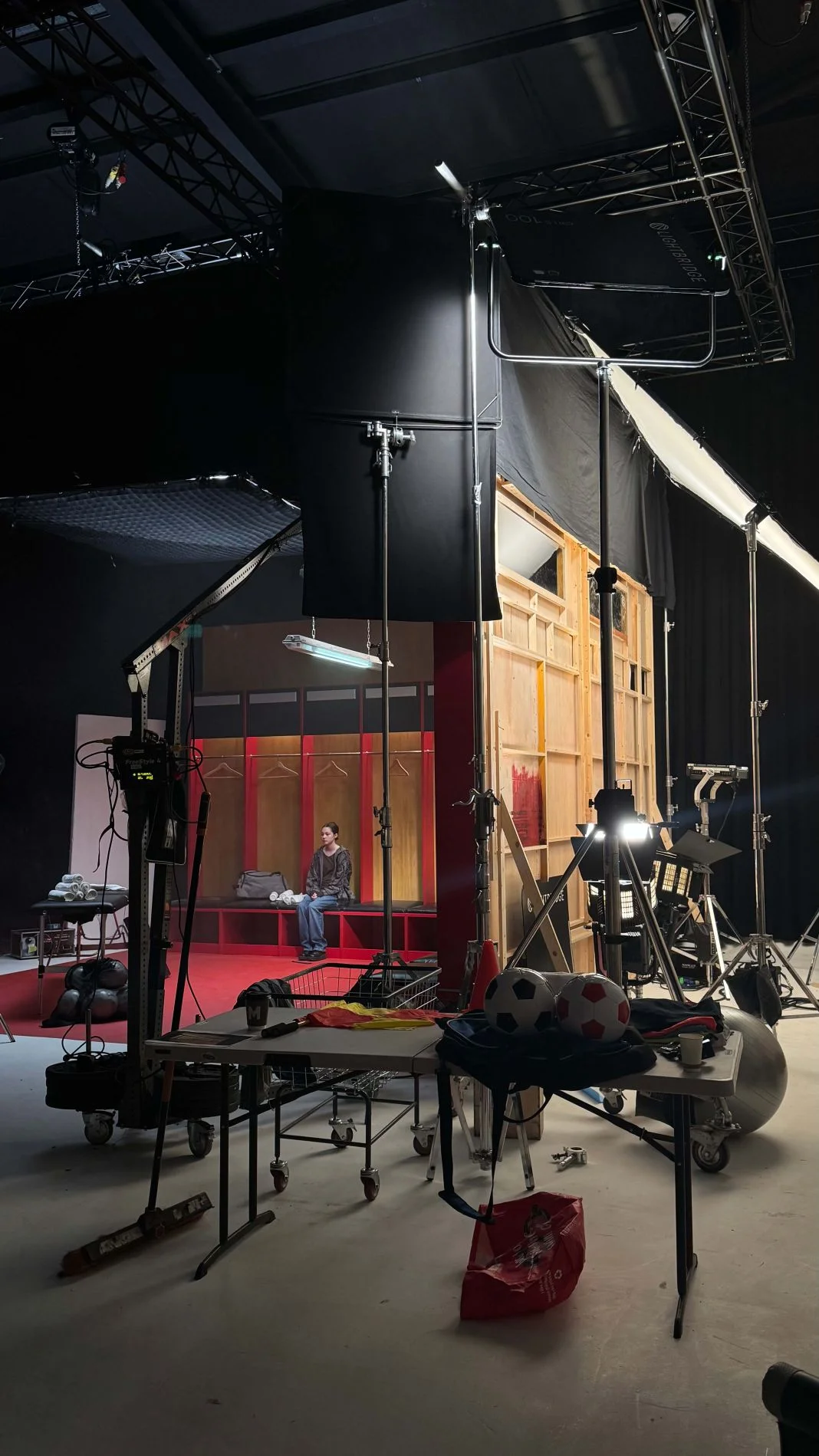 Film set with lighting rigs and equipment. A person sits on a red bench in a locker room. Soccer balls and gear visible.