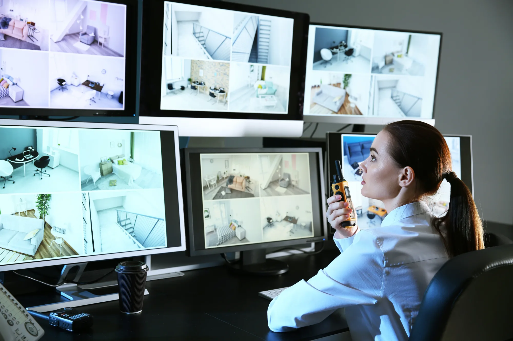 Woman in a white shirt monitors multiple security screens while holding a walkie-talkie, focused in a dimly lit control room.