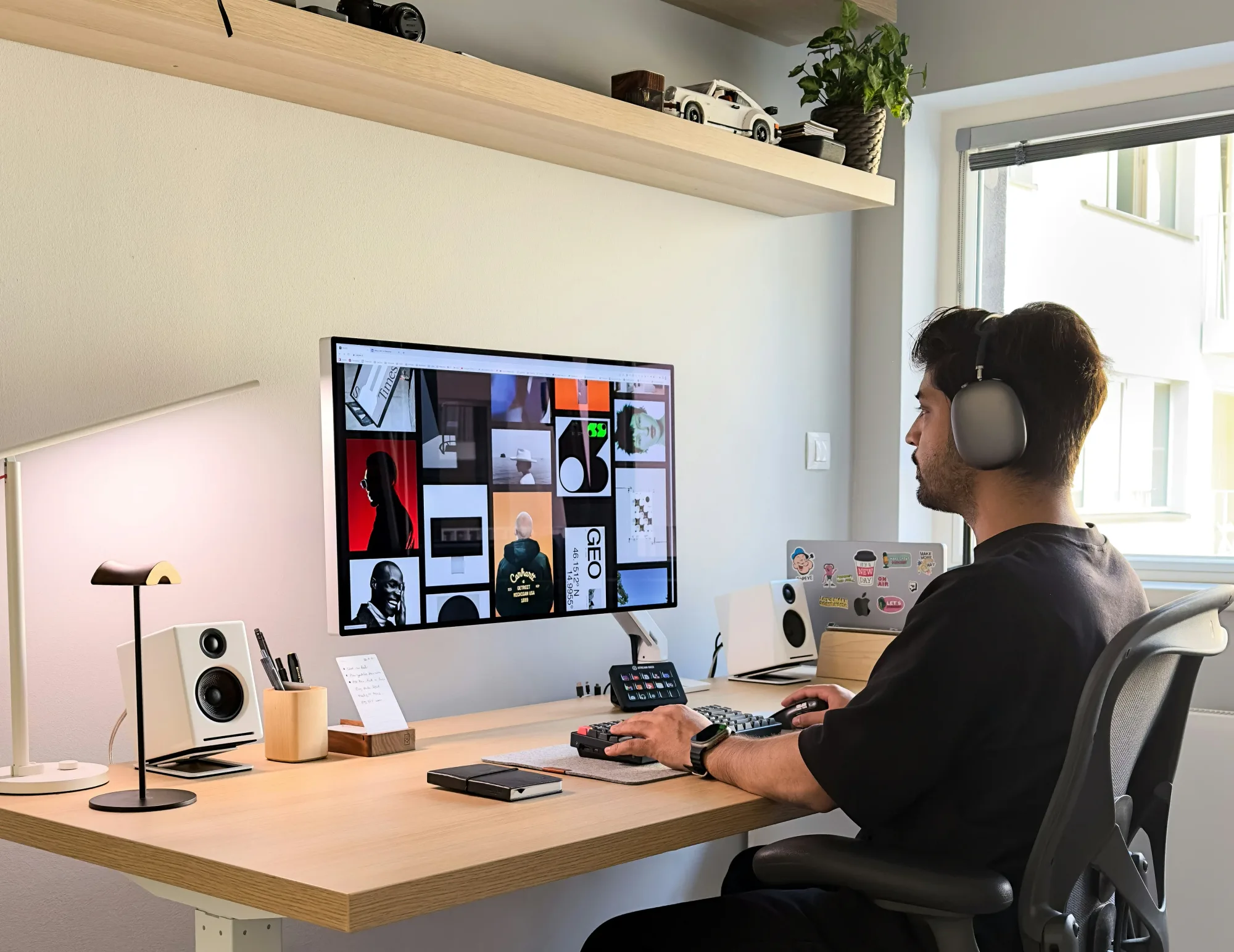 Man wearing headphones works at a desk with a large monitor displaying various images. Modern office setup with speakers and plants. Bright ambiance.