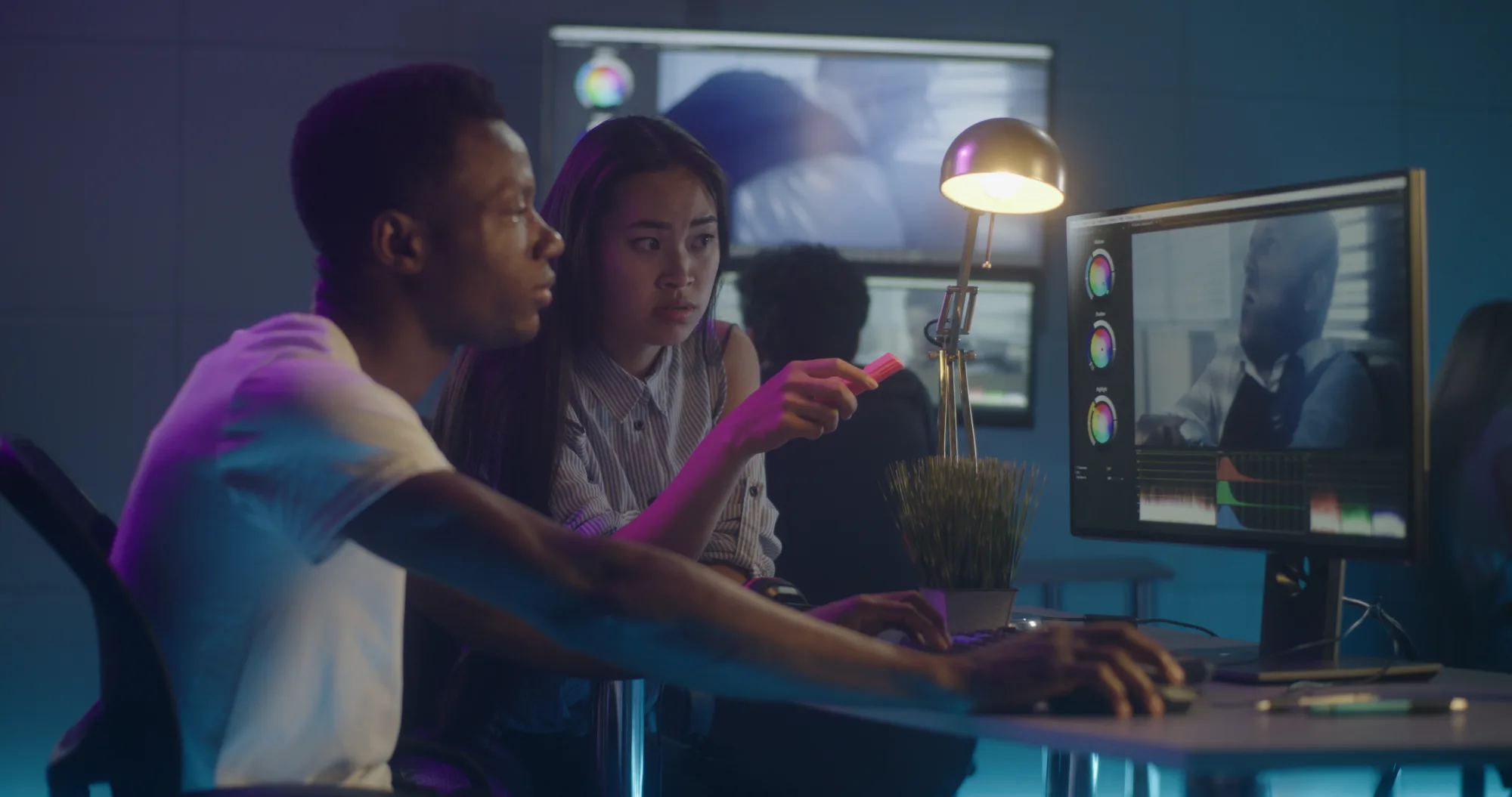 Two people focus on video editing at a computer in a dim room with blue lighting. The woman points at the screen displaying color wheels.