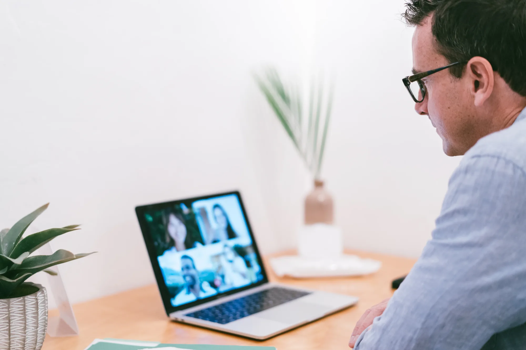 Man in glasses on video call with four people on laptop. Sitting at desk with plant, vase, and papers. White wall background.