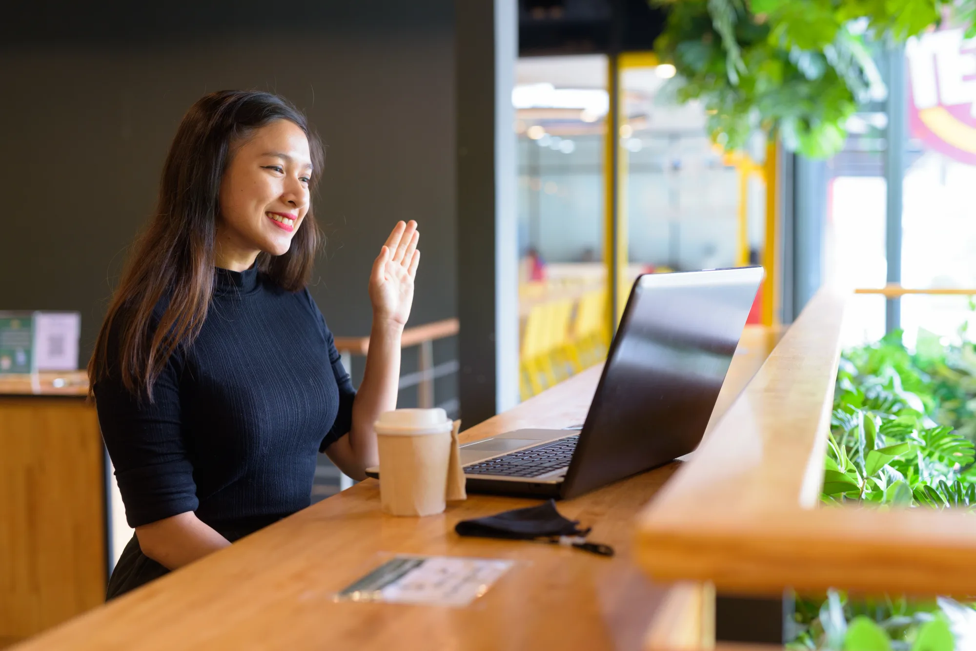 Woman in black top waves at laptop in a bright café; wooden counter, coffee cup, plants, and large windows create a cheerful vibe.