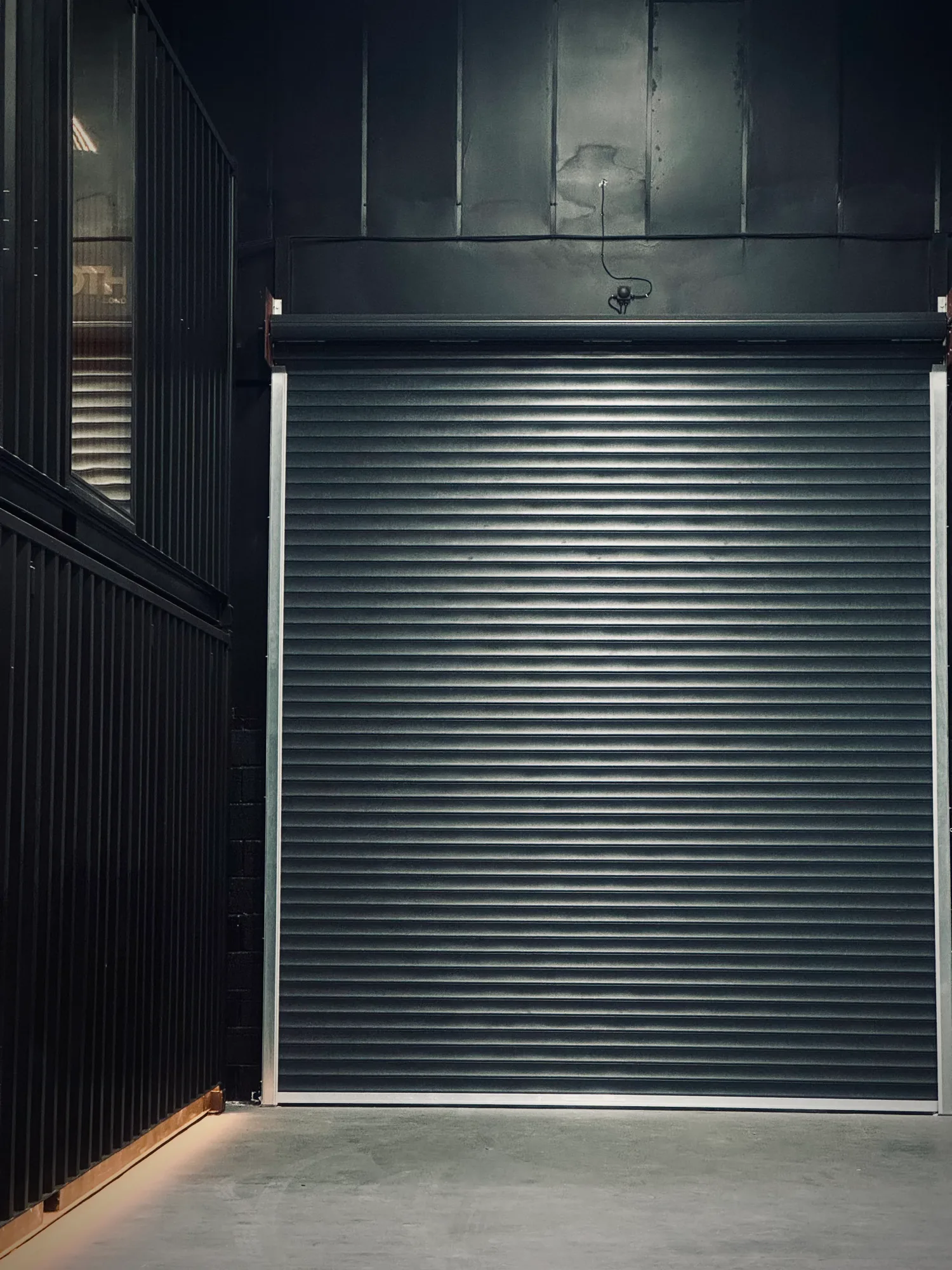 A closed, dark gray metal shutter door in an industrial setting with corrugated metal walls, creating a minimalist, somber mood.
