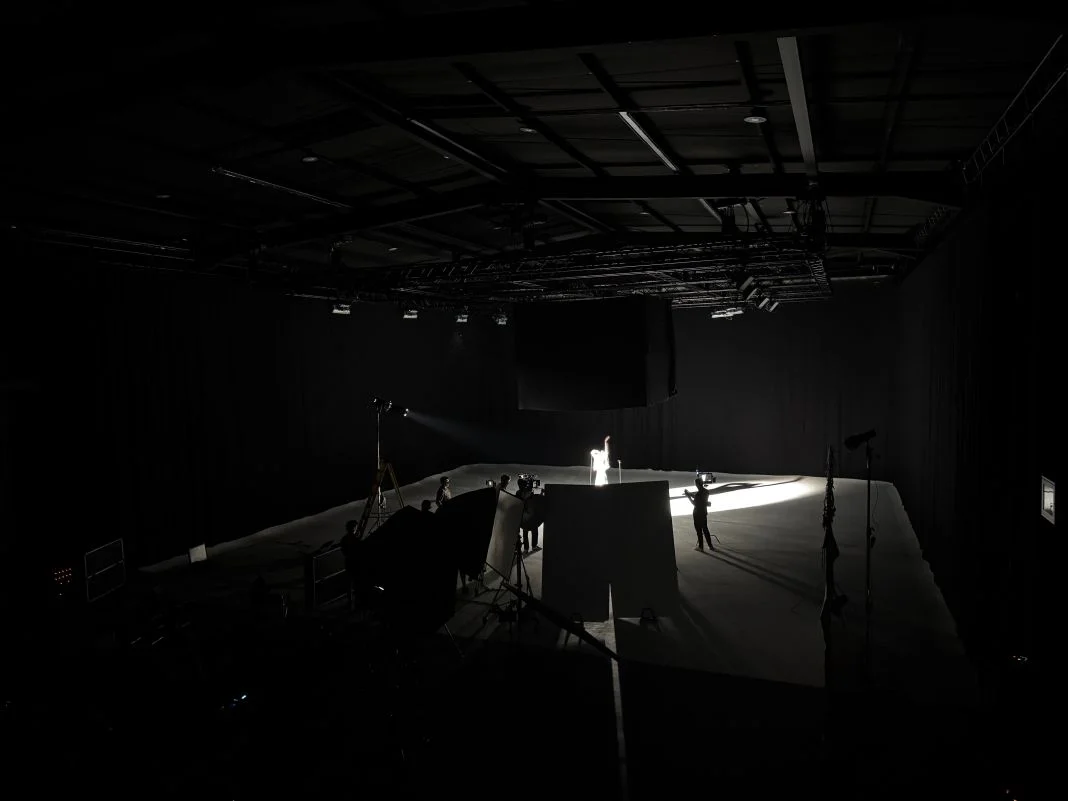 A person stands in a spotlight on a dimly lit stage, surrounded by crew members. The dark room creates a dramatic, focused atmosphere.
