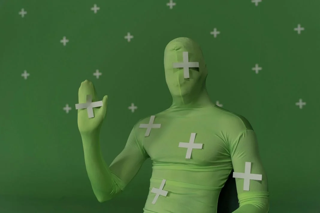Person in a green suit with plus signs, waving. Green background with white pluses. Neutral expression, surreal setting.