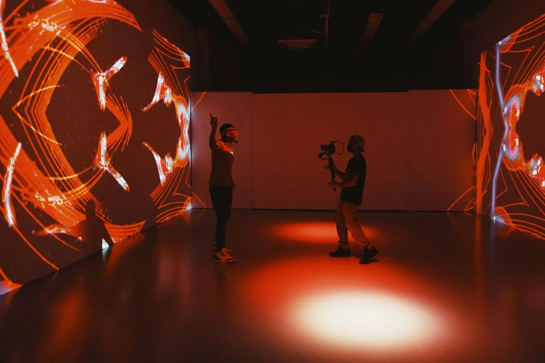Two people in a dim room, one filming, the other gesturing. Vivid red patterns are projected on surrounding walls, creating an artistic vibe.
