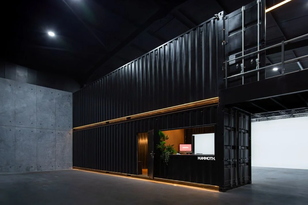 Black industrial space with a two-story container structure. A red screen and plant are visible at a window with "MAMMOTH" text. Minimal design.