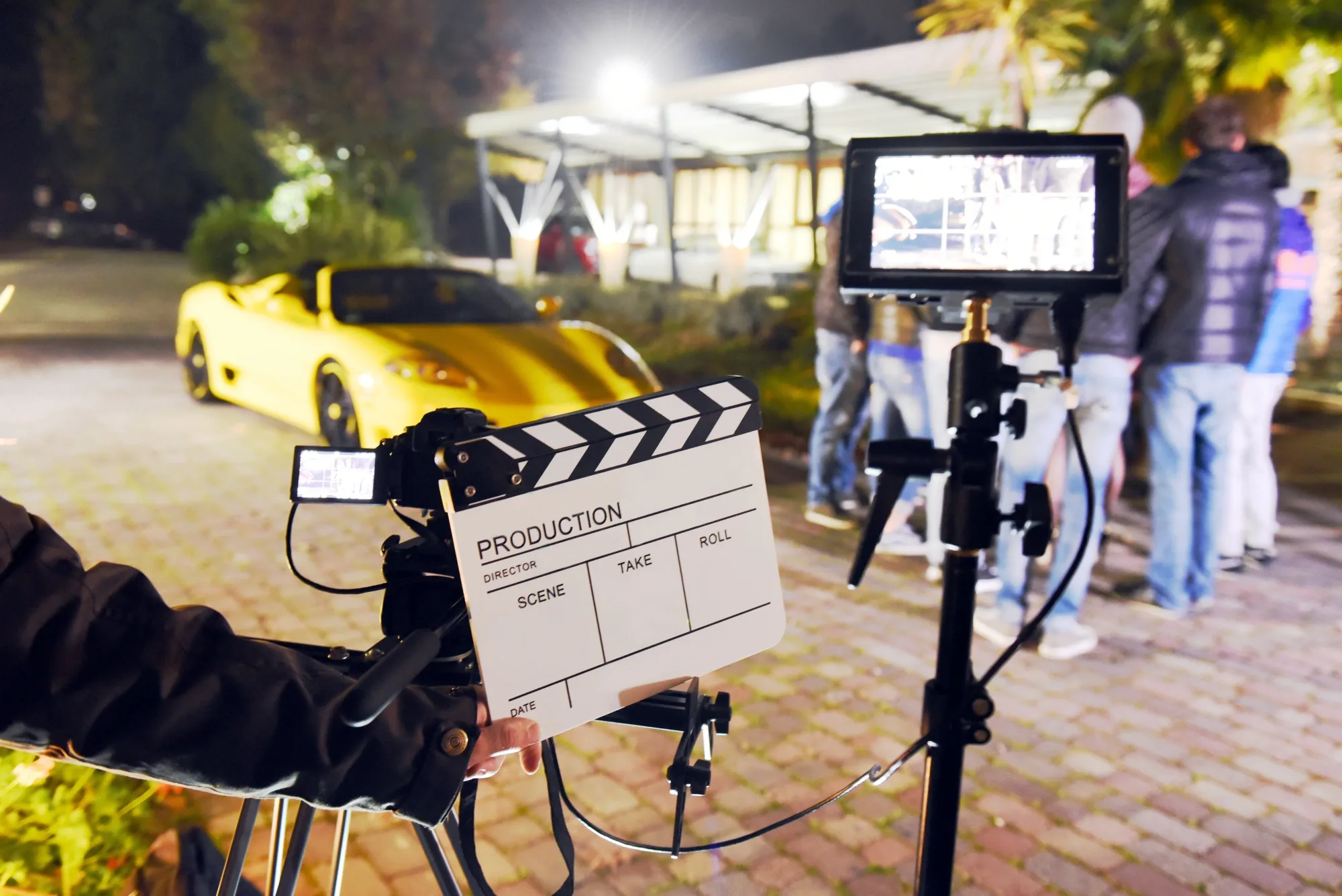 Film set at night with yellow sports car in background. Director holds clapboard near camera. Crew stands nearby, creating an active scene.