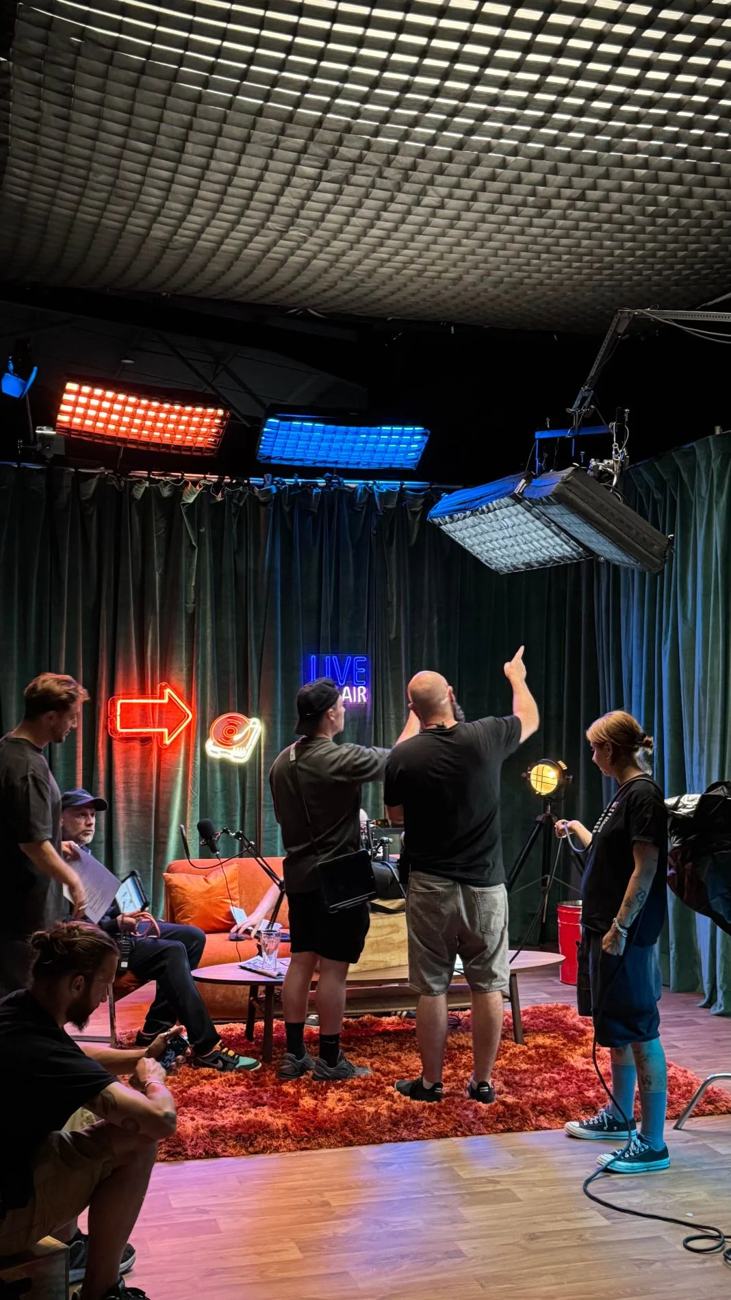Five people set up a studio with neon lights, a "LIVE ON AIR" sign, and equipment. The room is colorful and busy with a creative vibe.