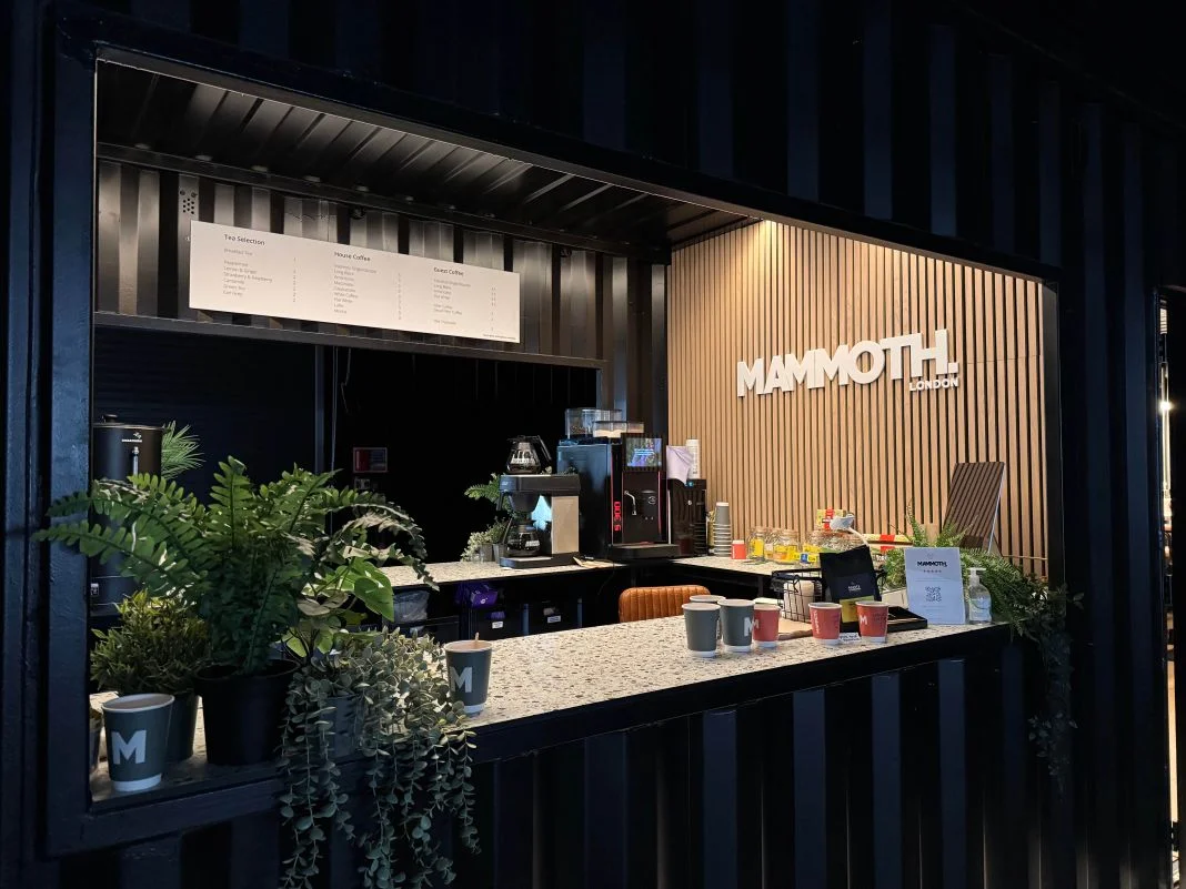 Coffee stand with plants, cups, and coffee machine. "MAMMOTH" sign on wooden wall. Menu above; cozy and inviting atmosphere.