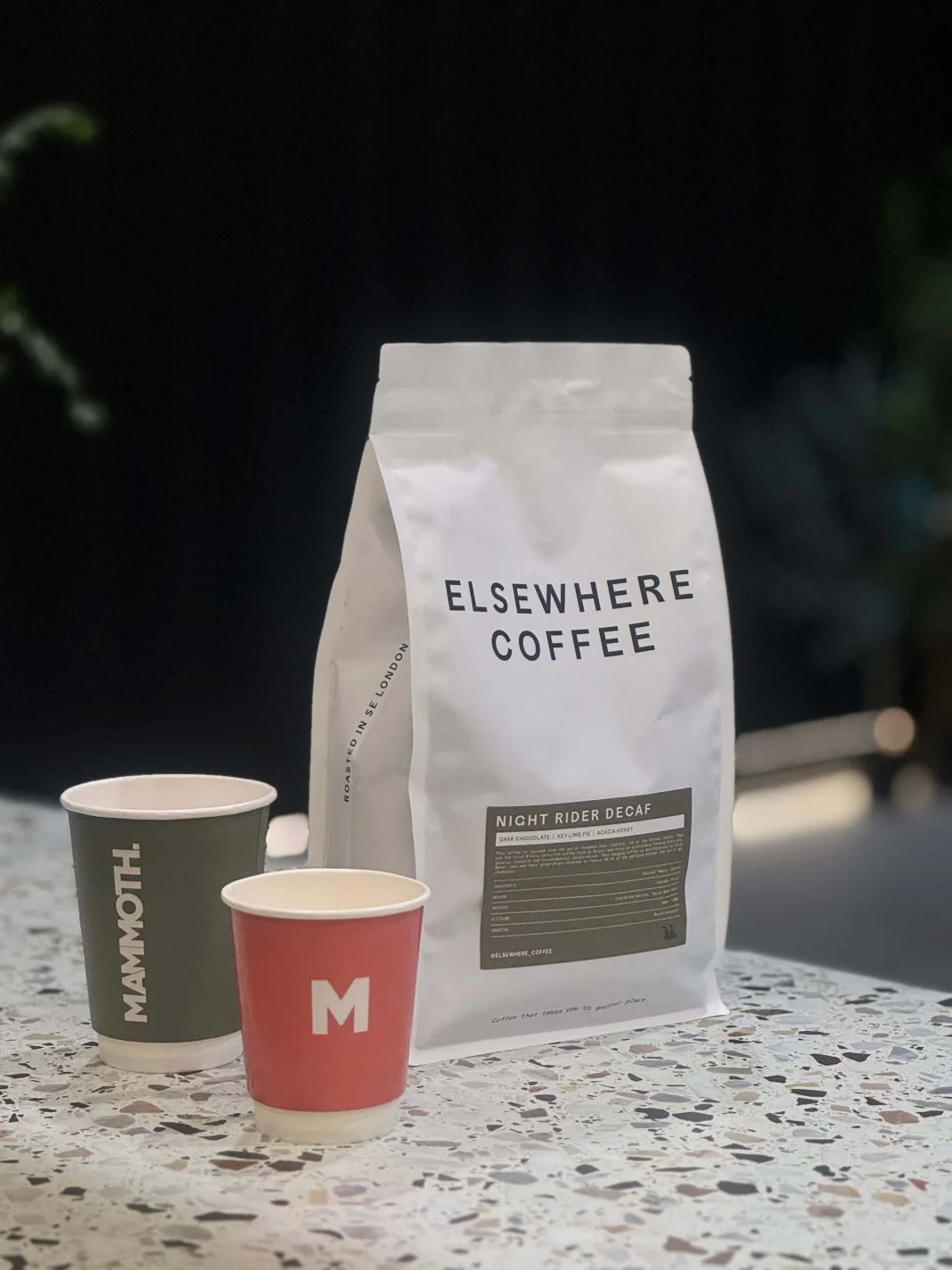 Bag of Elsewhere Coffee and two cups labeled Mammoth and M on a speckled table. The bag reads Night Rider Decaf. Dark background.