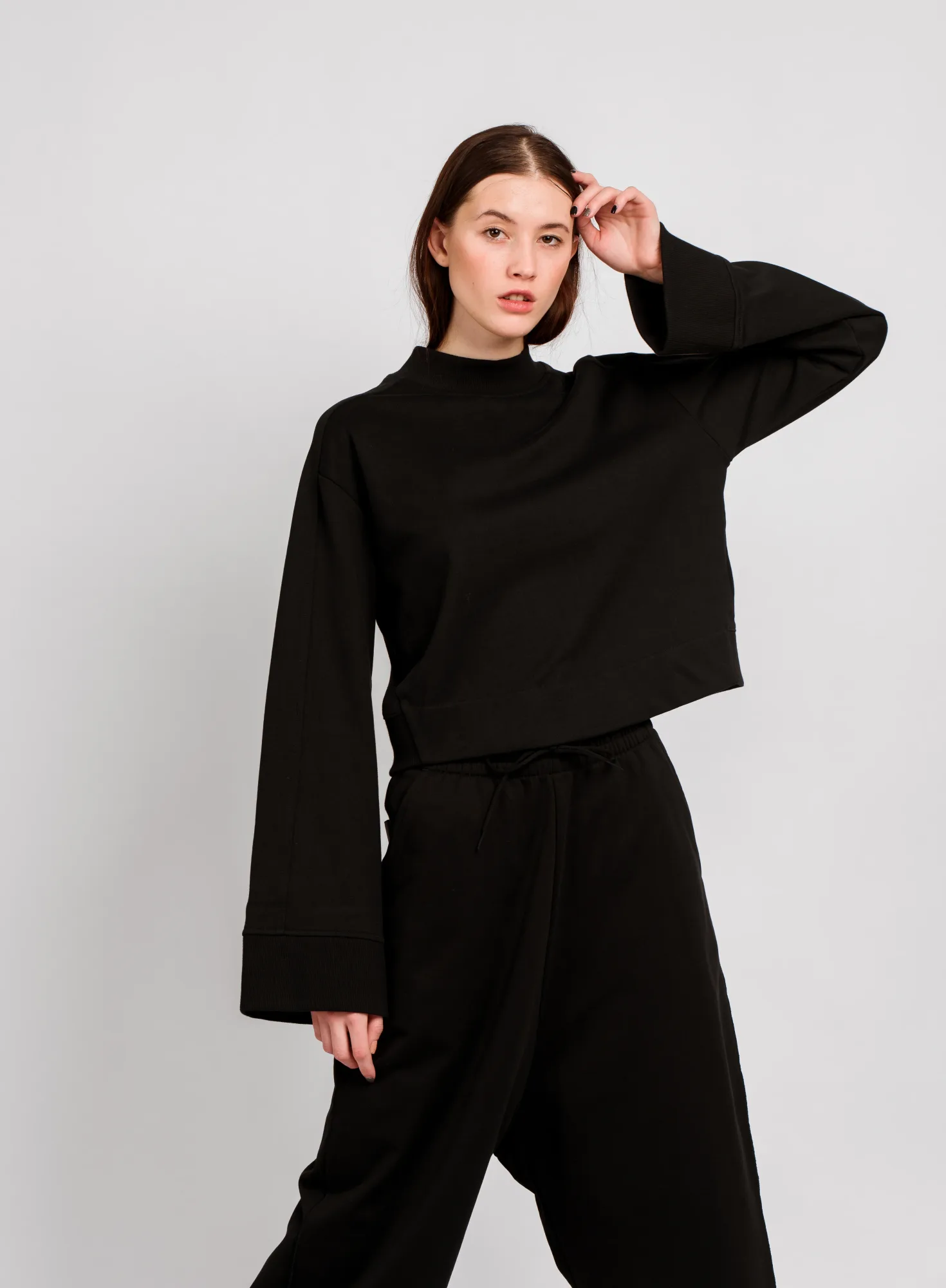 A person in a black outfit poses against a plain white background, looking directly at the camera with a neutral expression.