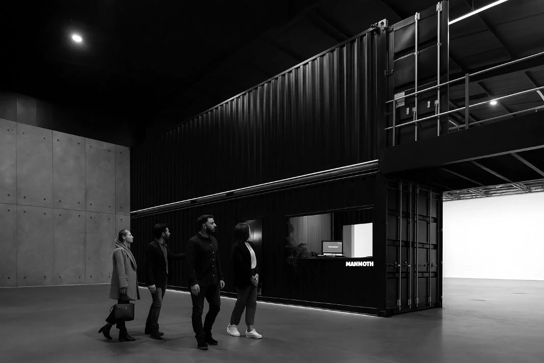 Four people walk past a black shipping container office in a large, industrial space. "MAMMOTH" is visible on the building. Monochrome theme.