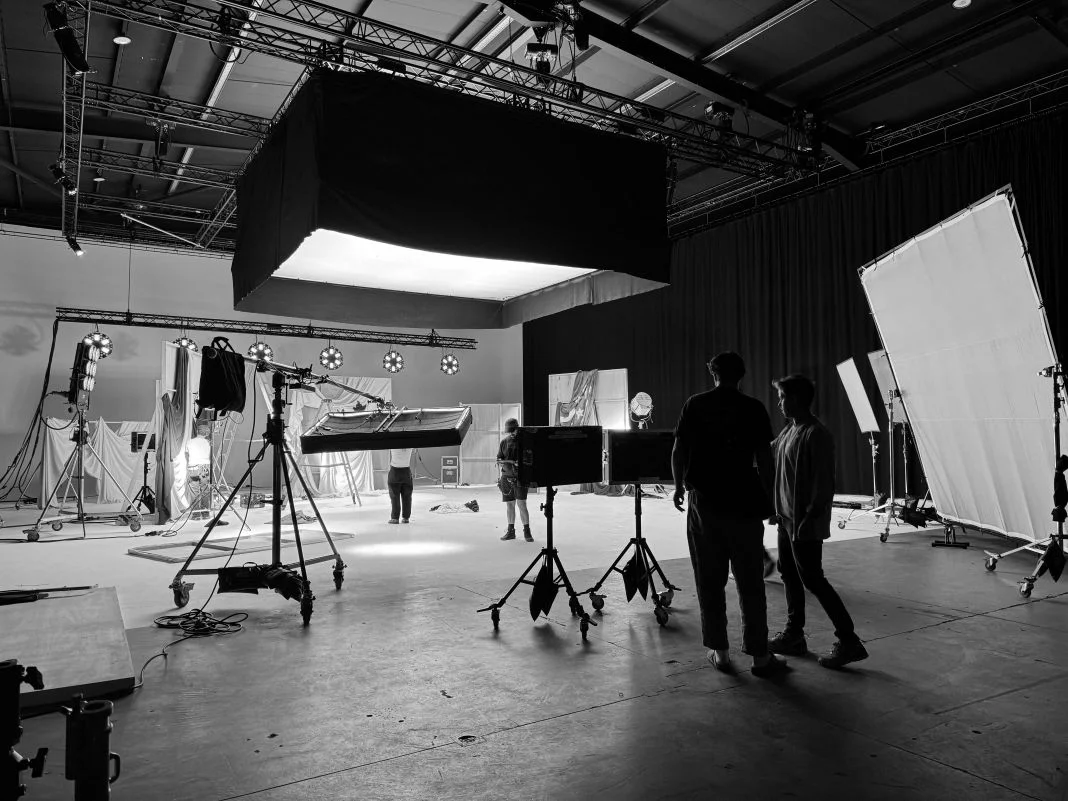 Film set in black and white, crew adjusts lights and equipment. Large lighting rigs create a focused, collaborative atmosphere.