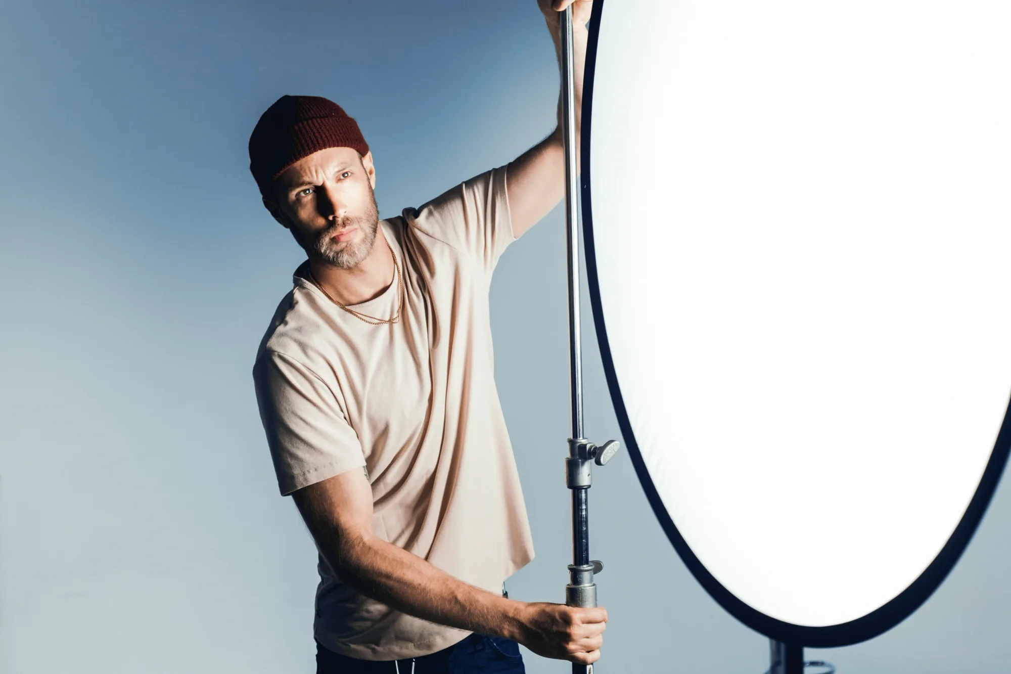 Man in a red beanie adjusts a large light reflector in a studio with a blue background. He wears a beige shirt, focusing intently.