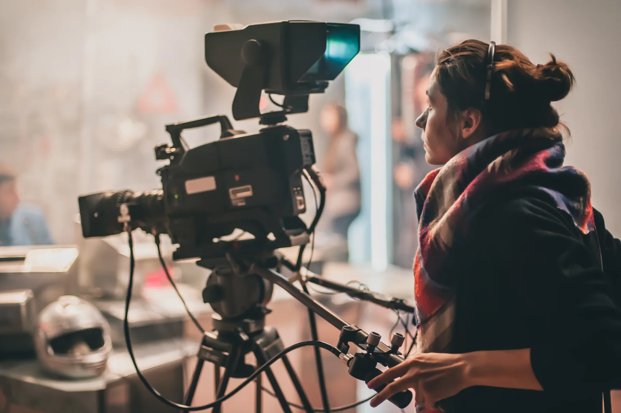 Camera operator focusing on a large video camera in a studio setting, wearing headphones. A blurred background and helmet are visible.