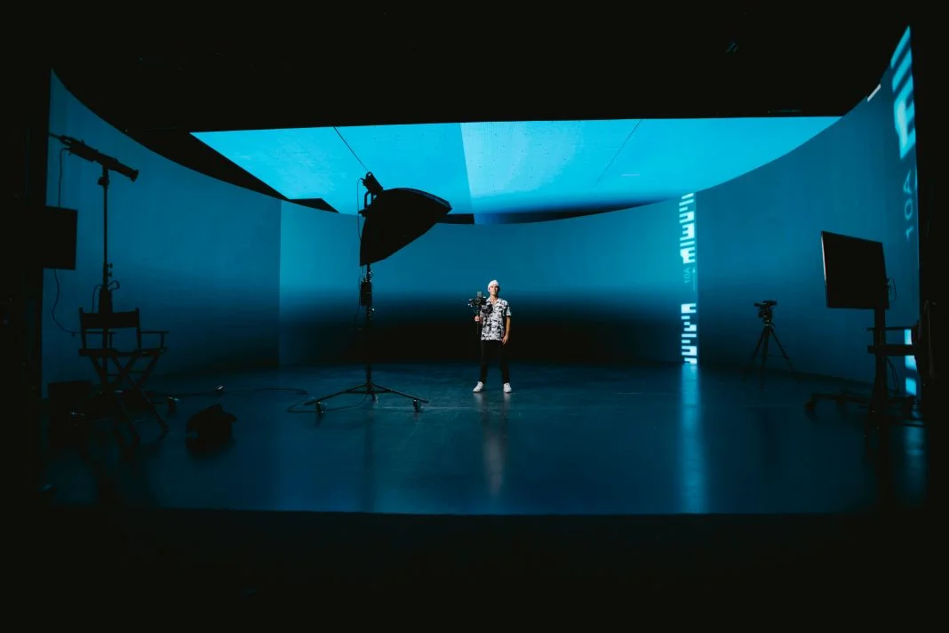A person stands in a blue-lit studio holding a camera. Surrounding are large screens, lighting equipment, and tripods. Calm atmosphere.