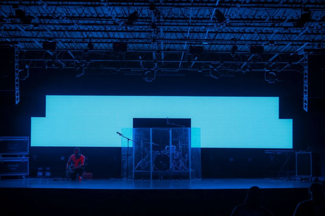 Stage with bright blue background, drums behind a transparent screen, person in red crouching on the left, microphone stand visible.