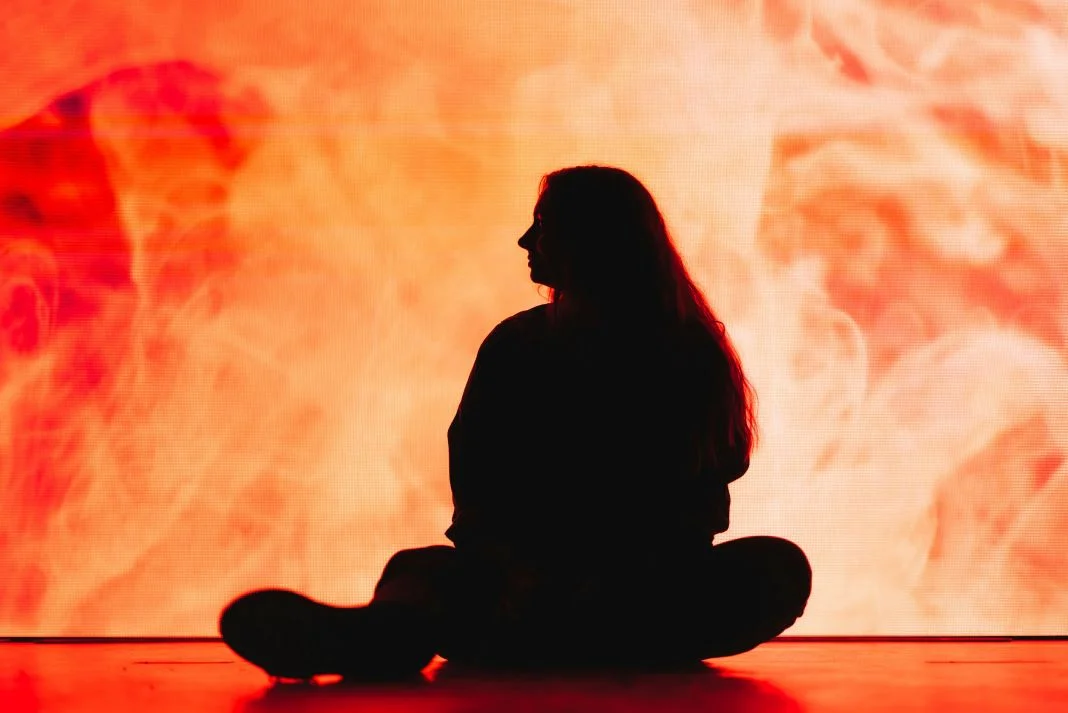 Silhouette of a person sitting cross-legged against an orange and red fiery background. The scene is dramatic and introspective.
