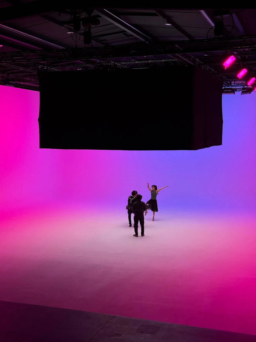 Dancer poses on one foot in a studio with pink and blue lighting, while two people film. The setting conveys creativity and energy.