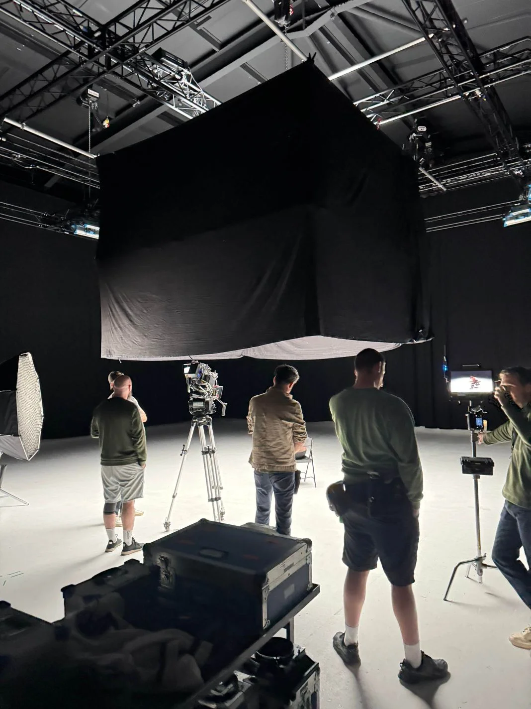 Film crew in a studio with camera equipment and lights, setting up a scene. The room has dark walls and a large black cloth overhead.