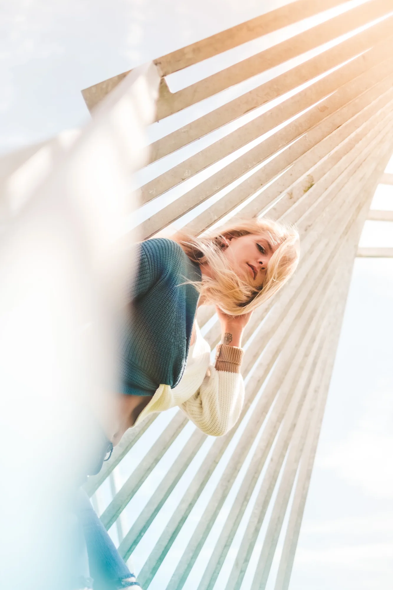 Woman with blonde hair poses beneath a geometric structure, wearing a blue and white top. Sunlight casts patterns, creating a dreamy vibe.