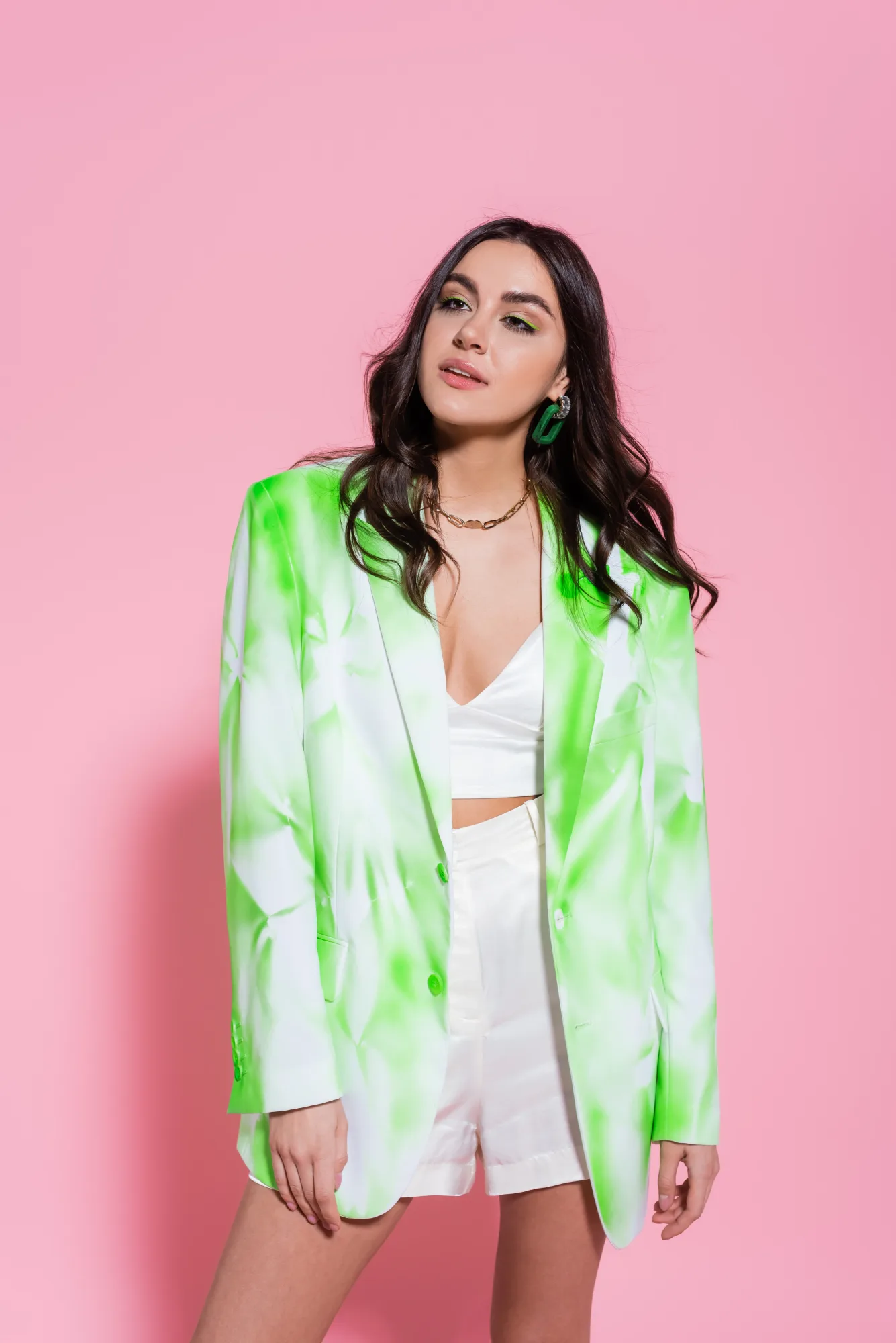 Woman in a white crop top and shorts, wearing a green patterned blazer. She's posed against a pink background, exuding a confident vibe.
