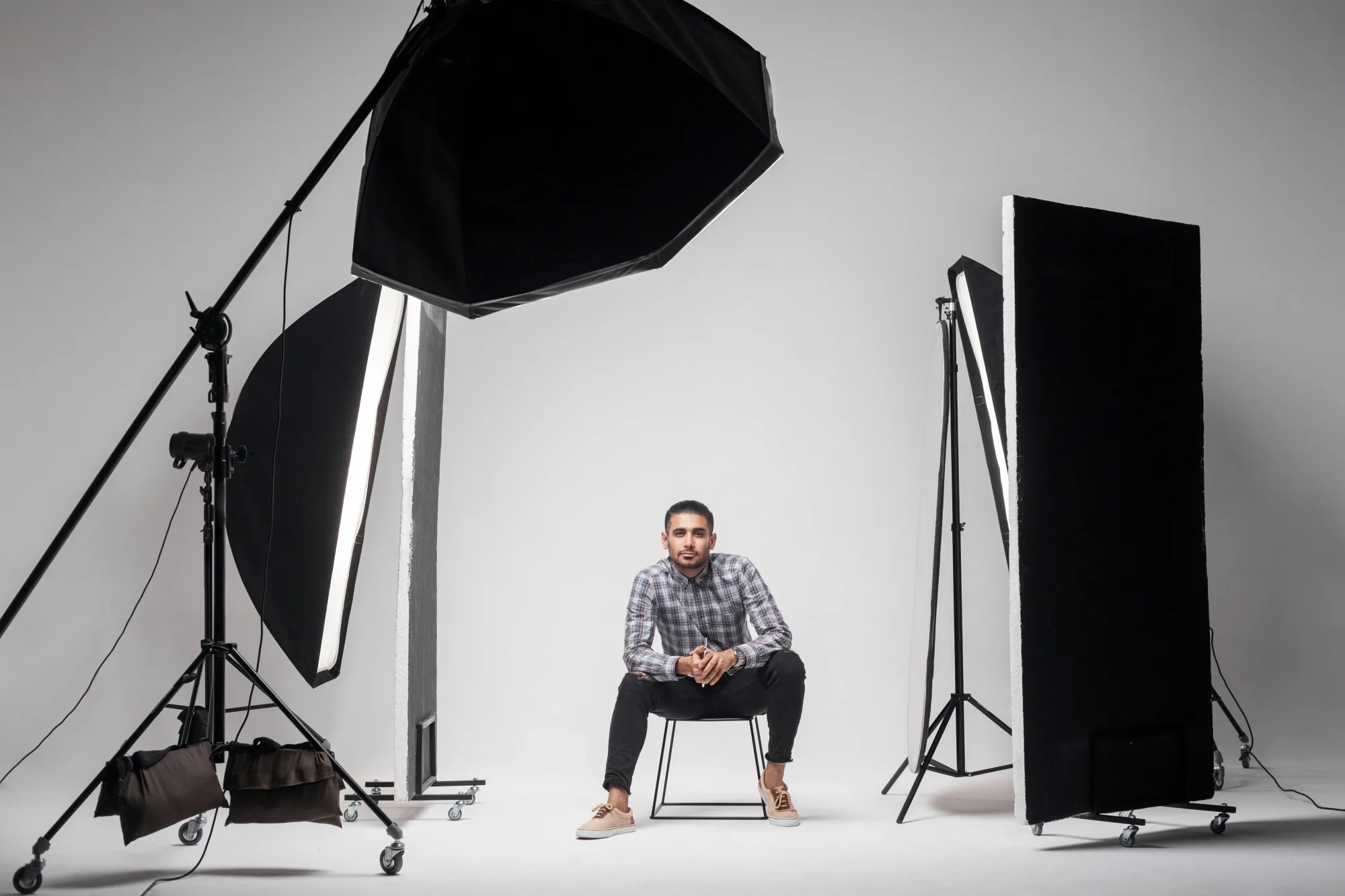Man in checked shirt sits on a chair in a photography studio, surrounded by large lighting equipment, against a plain white background.