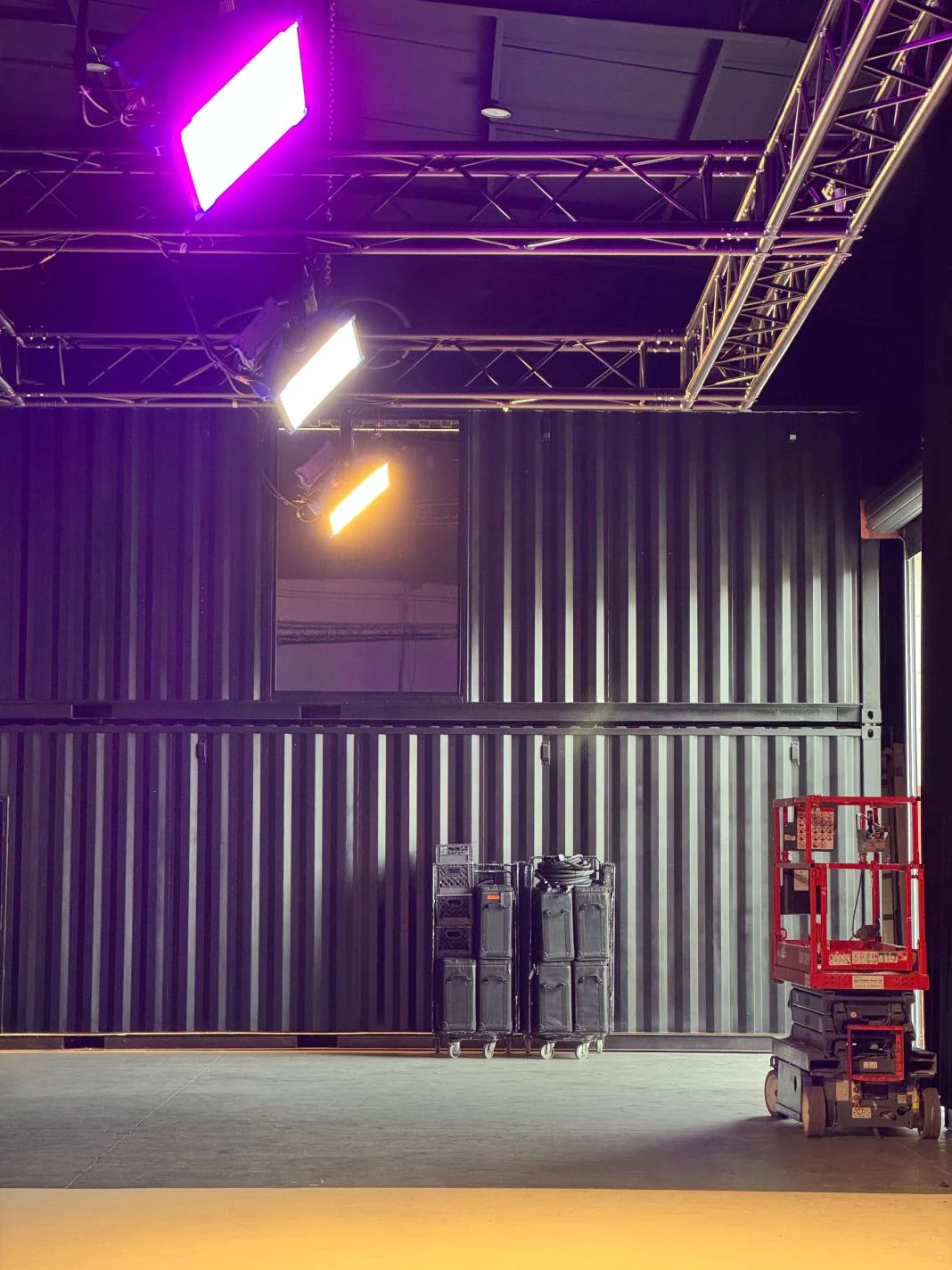 Stage setup with colored lights overhead, a red lift on the right, black equipment cases on the left. Industrial background, empty space.