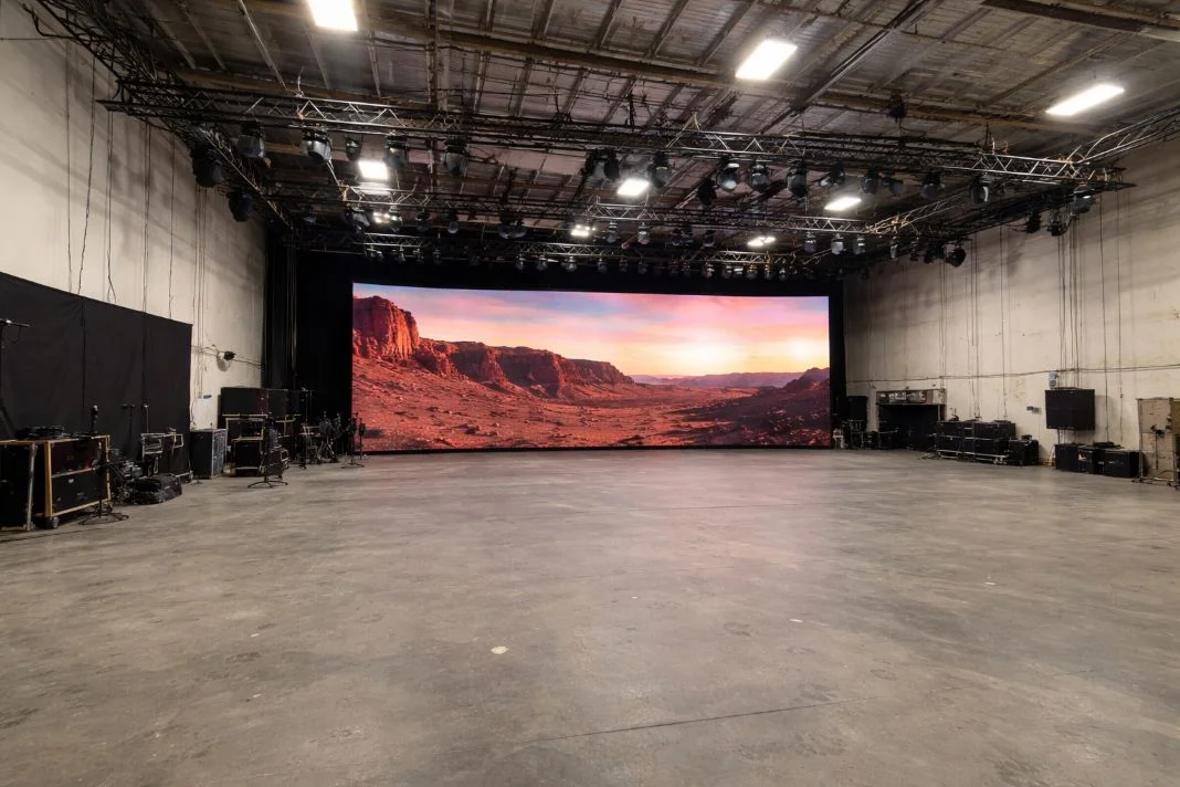 Large studio with a high ceiling displays a vivid desert landscape on a massive screen. Equipment lines the walls, creating an immersive setting.