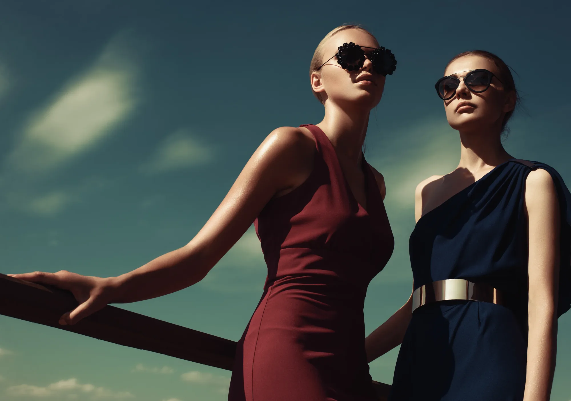 Two women in elegant dresses and sunglasses stand outdoors against a cloudy sky. One wears burgundy, the other navy with a gold belt.