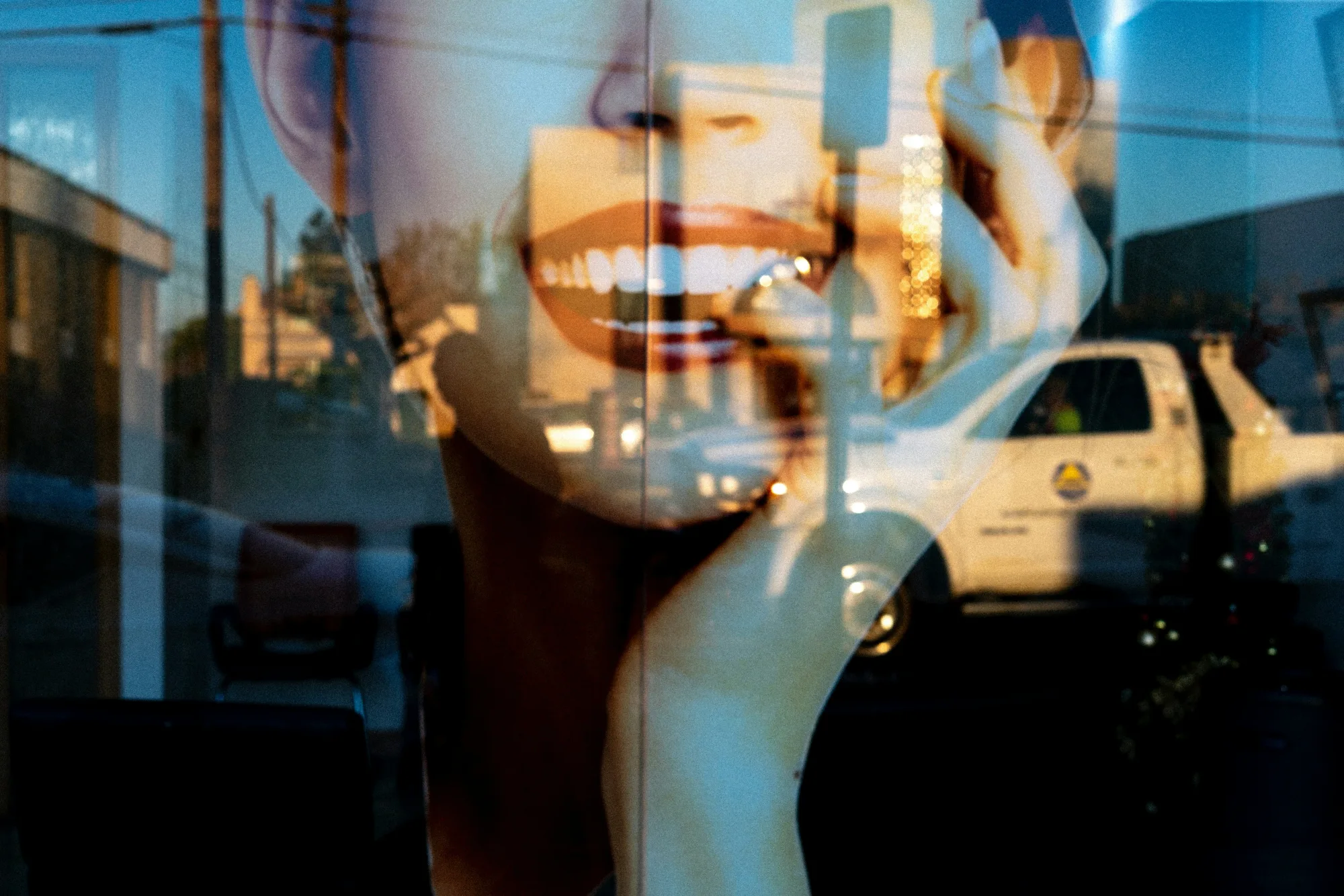 Reflection of a large smiling face on glass overlapped with outdoor street scene. Blue and orange tones. A parked white truck is visible.