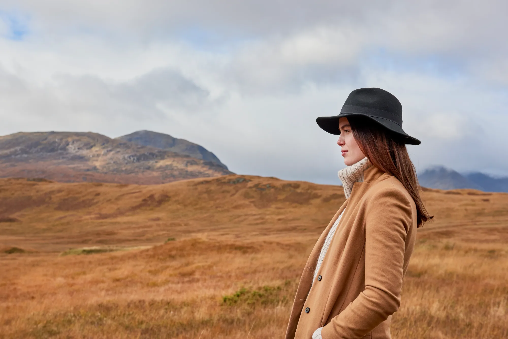 Woman in brown coat and black hat stands in a grassy field with mountains in the background. Overcast sky; calm mood.